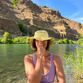Person wearing a yellow hat and purple top by a river with mountains in the background