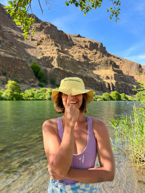 Person wearing a yellow hat and purple top by a river with mountains in the background